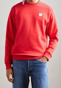 Sudadera roja con cuello redondo, puños acanalados y un pequeño parche de logo en el pecho. Combinada con vaqueros azules, presenta una textura suave.