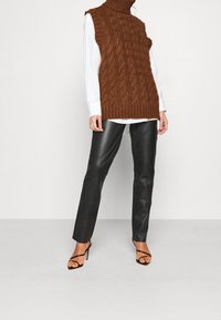 Hnědý pletený bezrukávový rolák s copánkovým vzorem, kombinovaný s černými koženými kalhotami a černými sandály s pásky a vysokými podpatky.