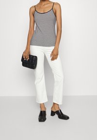 Mujer vestida con una blusa sin mangas a rayas en blanco y negro, pantalones blancos, mules negros con tacón, sosteniendo un bolso de mano acolchado negro.