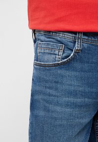 Chemise rouge rentrée dans un jean en denim bleu montrant des coutures détaillées et une poche avant sur un fond clair.