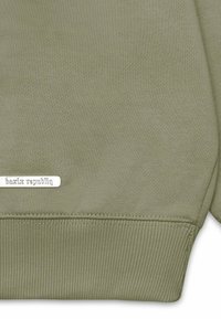 Olivgrüner Sweatshirt aus weichem Baumwollstoff. Mit geripptem Saum und Bündchen, versehen mit einem silbernen Etikett mit der Aufschrift "bazix republiq" am unteren Rand.