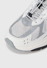 Sportowe sneakersy z siateczkową cholewką w jasnoszarym i białym kolorze, z gładkimi szarymi akcentami, sznurowadłami oraz teksturowaną podeszwą dla lepszej przyczepności.