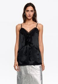 Czarna satynowa koszulka na cienkich ramiączkach z koronkow�ą lamówką, regulowanymi ramiączkami typu spaghetti i detalem wiązania z przodu. Noszona w połączeniu z metaliczną srebrną spódnicą.