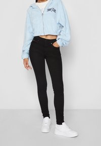 Sudadera corta con cremallera en azul claro y cintura elástica, jeans ajustados negros y zapatillas blancas. Textura suave y detalle de logotipo simple en la sudadera.