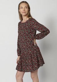 Rotes Blumenkleid mit langen Ärmeln, rundem Ausschnitt und gerafftem Rüschensaum. Der Stoff hat einen schwarzen Hintergrund mit mehrfarbigen Blumenmustern.