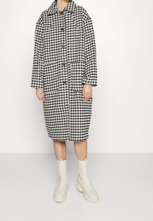 Personne portant un manteau mi-long à motif pied-de-poule noir et blanc avec des boutons noirs et des bottines blanches, debout devant un arrière-plan uni.