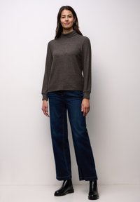 Donna in piedi contro una parete bianca che indossa un maglione marrone a collo alto, jeans blu scuro e stivaletti neri, rivolta in avanti con una postura rilassata.