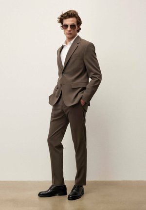 Vīrietis brūnā uzvalkā, baltā kreklā, melnās lakādas kurpēs un saulesbrillēs, stāv ar rokām kabatās pret vienkāršu fonu.