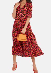 Robe midi fleurie avec un col en V, fond noir, fleurs rouges et orange, manches bouffantes et jupe à volants. Associée à un sac à main orange.