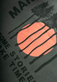 Svart t-shirt med en grafisk design som har en stor korallsol och mörka trädsilhuetter, kompletterad med texten "GÖR NÅGRA BERÄTTELSER TA TID."