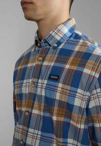 Hombre vistiendo una camisa de cuadros azul, naranja y blanca, con un pequeño parche de logo negro en el pecho, contra un fondo gris.