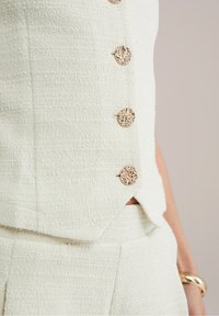 Cremfärgad texturerad väst med dekorativa guldbanor, med en skräddarsydd design och subtil slits på sidan. Slät tyg med ett vävt mönster.