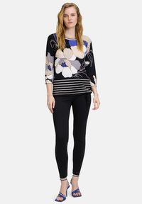 Langärmeliger Blumenpullover in Schwarz, Creme und Blau; gestreifter Saum. Figurbetonte schwarze Leggings kombiniert mit blauen Absatzsandalen.