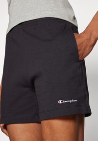 Pantalones cortos de algodón negros con bolsillos laterales, con un logo de Champion en blanco y rojo en el dobladillo. Textura suave y diseño de corte recto.