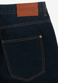 Granatowe dżinsy z denimu z skórzaną naszywką oznaczoną "PROSTO". Zawierają widoczne szwy i mały złoty nity na tylnej kieszeni.