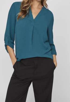 Vrouw draagt een tealblauwe blouse met lange mouwen en V-hals, met opgerolde mouwen en zwarte broek, handen in de zakken, tegen een effen achtergrond.