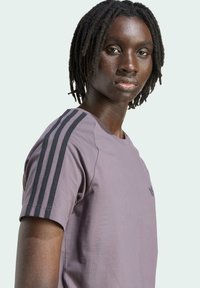 Camiseta atlética púrpura con tres franjas negras en los hombros, fabricada en tela suave con cuello redondo y un logo minimalista.