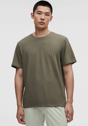 Ung mand stående iført en olivengrøn kortærmet t-shirt og lyse bukser mod en ensfarvet lys baggrund.