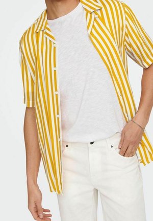 Mann mit einem gelb-weiß vertikal gestreiften Kurzarmhemd über einem weißen T-Shirt und weißen Hosen, eine Hand in der Hosentasche.