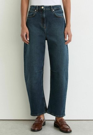 Persoon die donkerblauwe rechte jeans en bruine leren loafers draagt, staand tegen een witte muur en een grijze vloer.