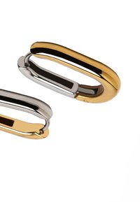 Dwie wypolerowane owalne kolczyki w kształcie obręczy, jedna złota, a druga srebrna, umieszczone na białym tle.