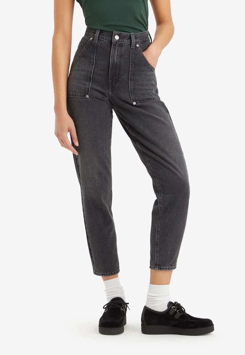 Högmidjade svarta denimjeans med avslappnad passform, utrustade med framfickor och silverdetaljer. Kombinationen med svarta skor och vita strumpor.