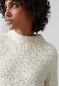Maglione texturizzato di colore crema con colletto alto e un aspetto morbido, caratterizzato da una vestibilità ampia e dettagli di cucitura sottili.