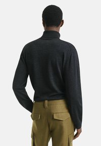 Donkergrijze langemouwen coltrui met textuur breiwerk, gecombineerd met olijfgroene cargo-stijl broeken met achterzakken en knoopdetails.