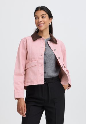 Femme souriante portant une veste rose avec un col marron par-dessus un pull gris boutonné et un pantalon noir, posant avec une main dans la poche.