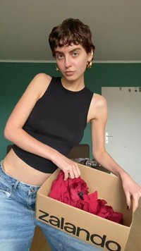 Juoda be rankovių palaidinė, šviesiai mėlyni džinsai ir laikantis Zalando dėžę, pilną raudonų audinių. Paprasta aplinka su žalia siena.