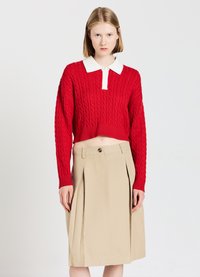 Roter, kürzer geschnittener Pullover mit Kabelstrickmuster und weißem Kragen, kombiniert mit einem beigen Faltenrock. Das Outfit zeichnet sich durch texturierte Materialien und kontrastierende Farben aus.