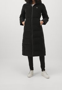 Kvinna klädd i en lång svart vadderad jacka med dragkedja framtill, händerna i fickorna, i kombination med svarta byxor och vita snörskor.