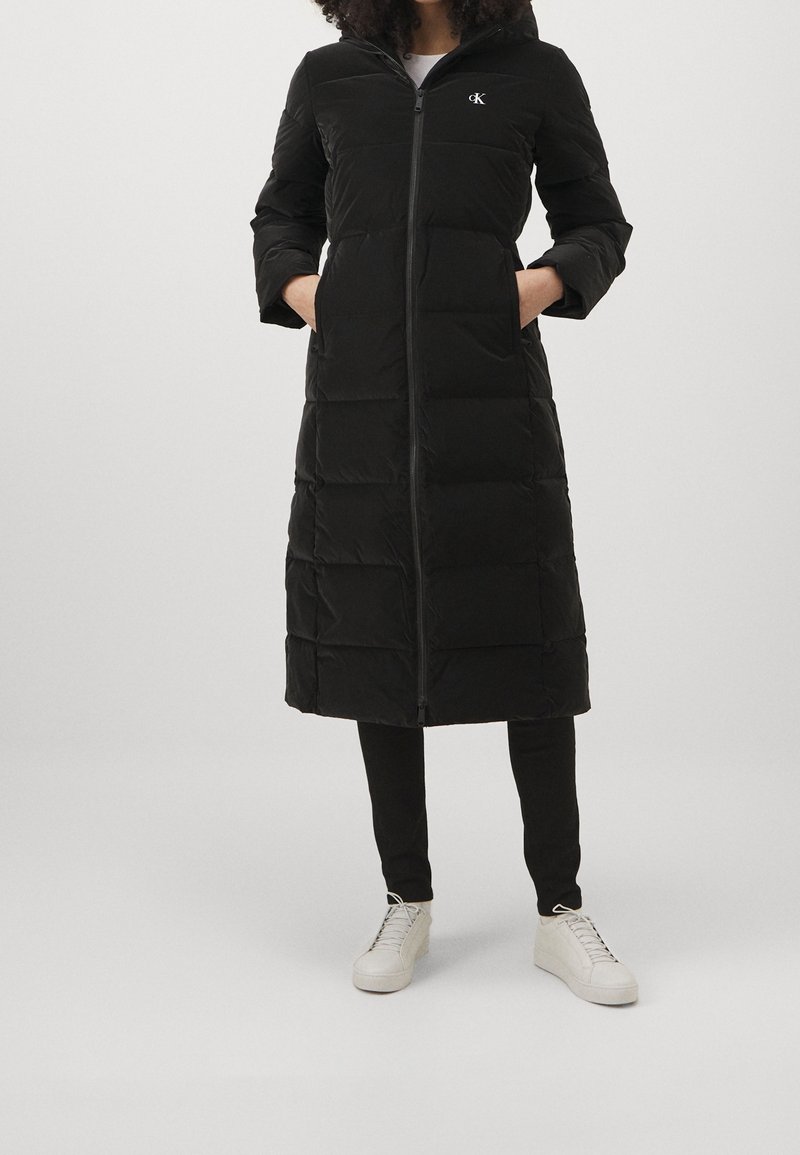 Kvinna klädd i en lång svart vadderad jacka med dragkedja framtill, händerna i fickorna, i kombination med svarta byxor och vita snörskor.