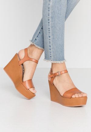 Brązowe sandały na koturnie ze gładką skórzaną cholewką, szerokimi paskami i zapięciem na kostkę. Platforma z delikatnym połyskiem. Jeansy z falbaniastym dołem.