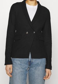 Černý blazer s texturovanou tkaninou, s šálovým límcem, zapínáním na dvojité knoflíky a dvěma předními kapsami, kombinovaný s modrými džíny.