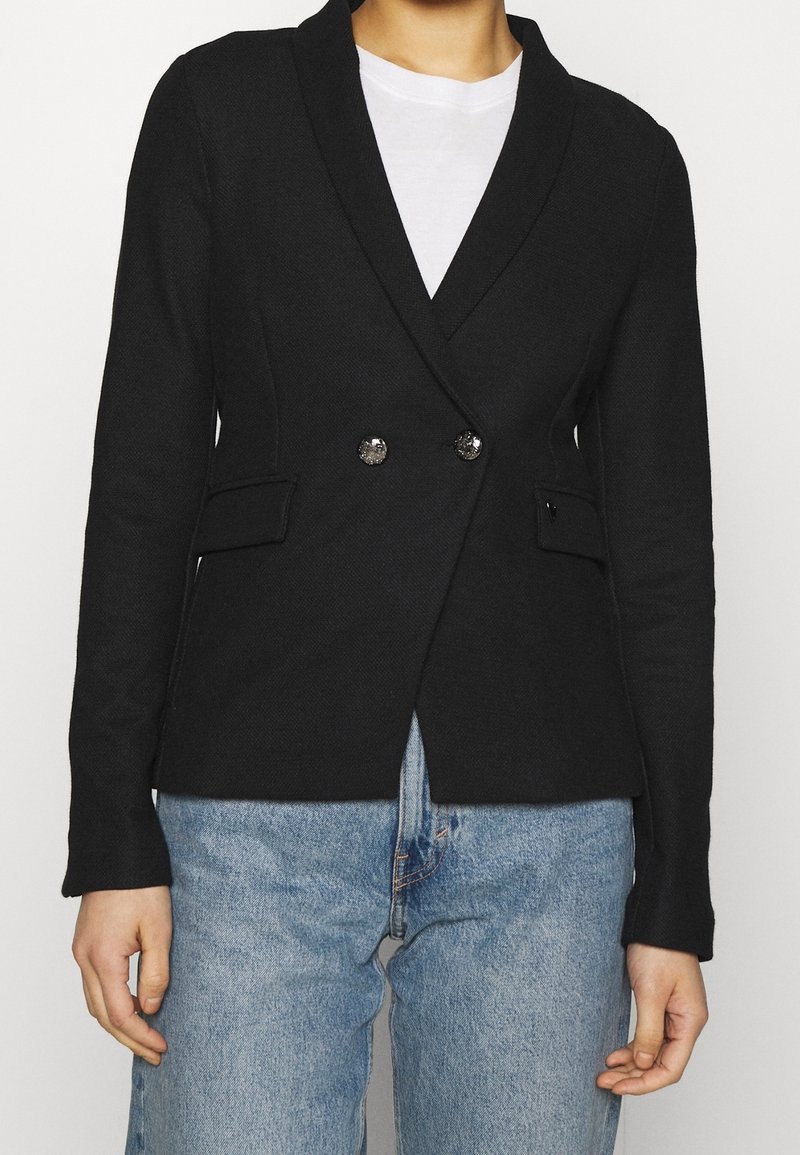 Černý blazer s texturovanou tkaninou, s šálovým límcem, zapínáním na dvojité knoflíky a dvěma předními kapsami, kombinovaný s modrými džíny.