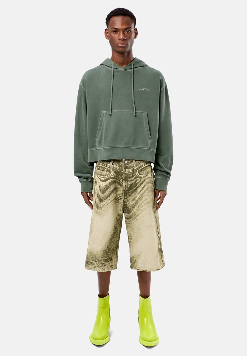 Felpa verde con cappuccio e lacci, abbinata a pantaloni corti metallizzati oro e stivali alla caviglia verde brillante. Superfici lisce con colori distintivi.