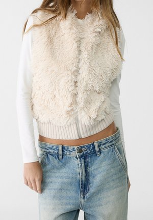 Femme portant un gilet zippé pelucheux couleur crème sur une chemise blanche à manches longues et un jean denim bleu clair ample.