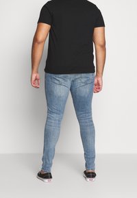 Hombre vestido con una camiseta negra, jeans de mezclilla ajustados de color azul claro y zapatillas negras, de pie con la espalda hacia la cámara frente a un fondo simple.