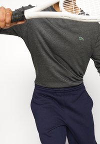 Gray long-sleeve shirt with a small green crocodile logo, paired with navy sweatpants. The shirt exhibits a smooth, soft texture.