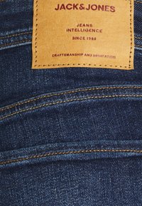 Vaqueros de mezclilla de color azul oscuro, con una etiqueta de cuero que dice "JACK & JONES", con costuras visibles y detalles de textura.