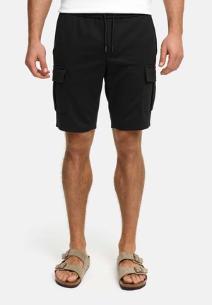 Mężczyzna ubrany w czarne krótkie spodnie typu cargo z bocznymi kieszeniami i beżowe sandały na podwójny pasek, stojący na tle białego tła.