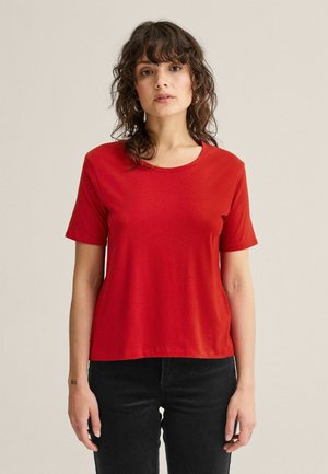 Kvinde med bølget mørkt hår iført en ensfarvet rød T-shirt med korte ærmer og sorte bukser, stående mod en neutral baggrund.