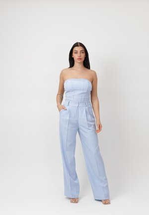 Donna con capelli scuri che indossa una tuta azzurra senza spalline e a gambe larghe, in piedi con una mano in tasca contro uno sfondo semplice.