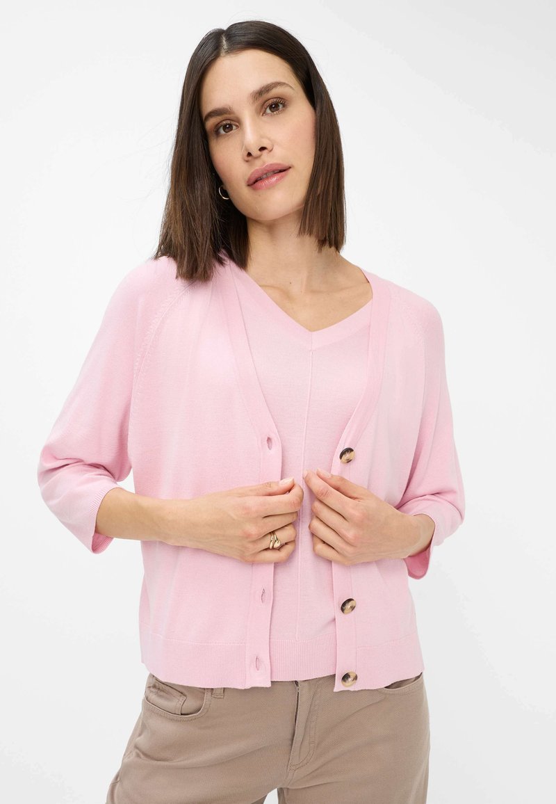 Light pink cardigan with three-quarter sleeves, button closure, and ribbed hem. Worn over a matching pink v-neck top. Smooth texture.