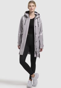 Geacă de ploaie waterproof, de culoare gri deschis, cu glugă, capse și accente cu șnur, purtată cu colanți negri și adidași gri.
