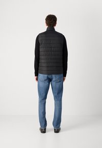 Colete acolchoado preto com costura horizontal, gola alta, usado sobre uma camisa preta de mangas longas, combinado com jeans azuis e sapatos pretos.