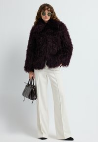 Kvinde står iført en mørk lilla, langhåret pelsjakke, hvide brede bukser, sorte sko, solbriller, guld hoops øreringe og holder en sort håndtaske.