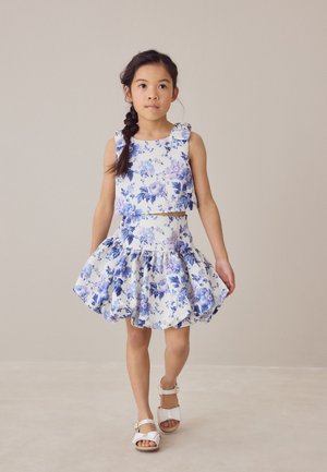 Chica con el pelo trenzado que lleva una blusa sin mangas con estampado floral blanco y azul y una falda acampanada a juego, caminando con sandalias blancas.
