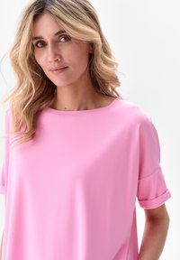 Jasnoróżowa bluzka z krótkim rękawem, z okrągłym dekoltem i podwiniętymi mankietami, wykonana z gładkiego, elastycznego materiału. Prosty i casualowy krój.